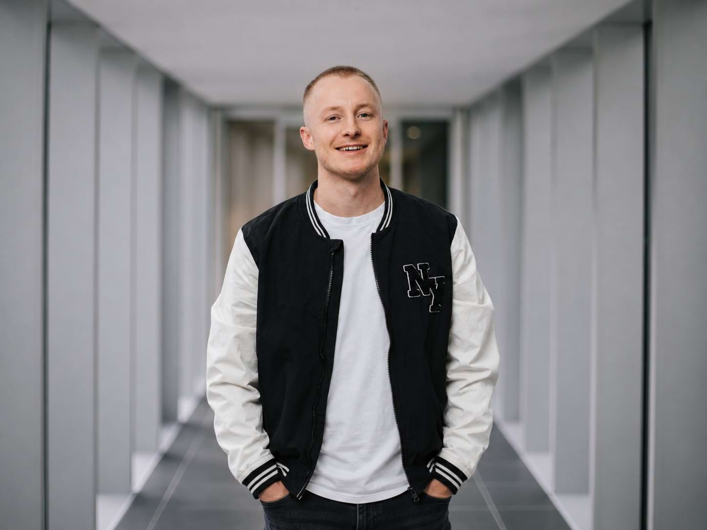 Mann mit kurzem Haar und Lächeln trägt eine schwarze und weiße Bomberjacke über einem weißen T-Shirt in einem modernen Innenr